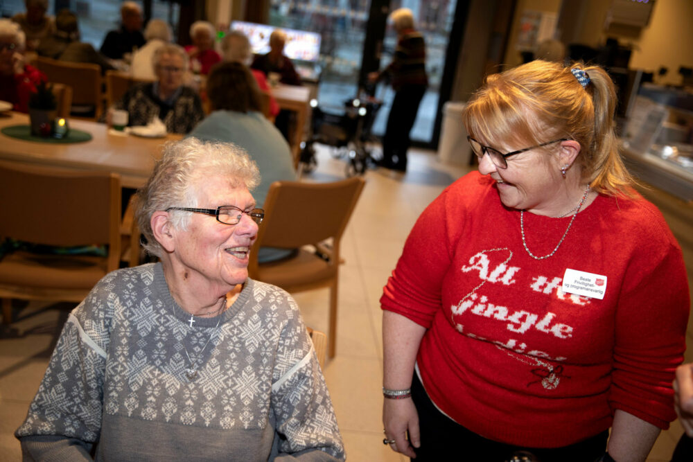 Foto: To damer snakker med hverandre, hun til venstre er eldst og sitter. Hun andre står og har på seg en rød julegenser. Begge har briller.