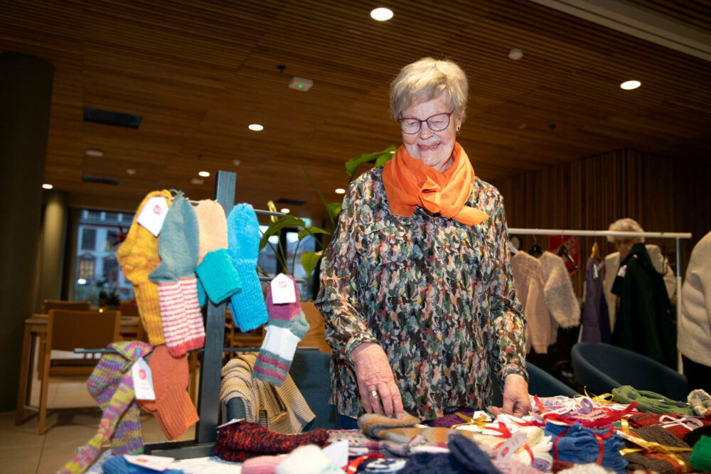 Foto: Eldre kvinne i 80-årene med kort, grått hår og briller, står ved et bord. her ligger det mange strikkede strømper og annet.