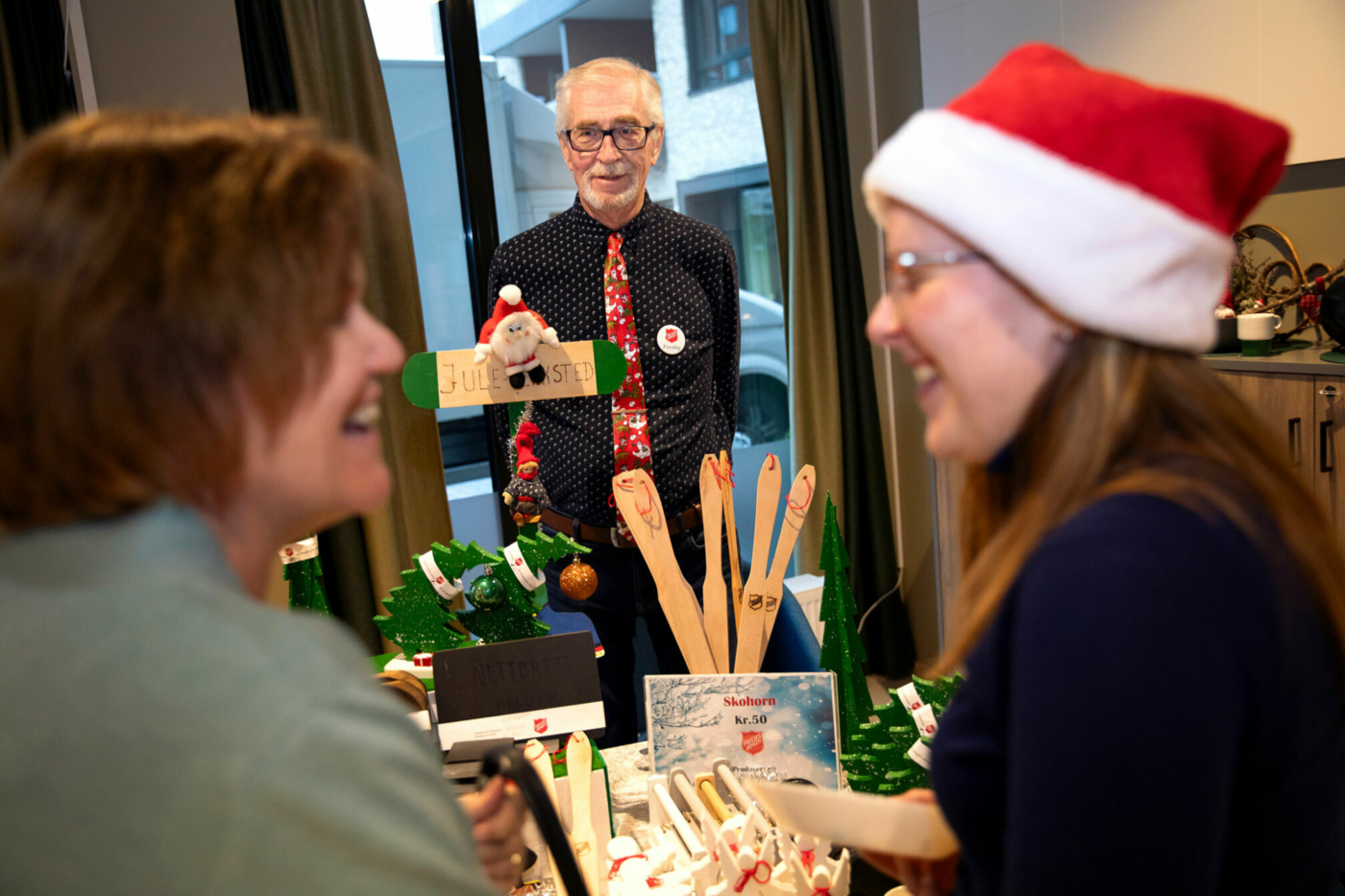 Foto: Pensjonist med rødt juleslips og briller står bak et bord med en rekke trevarer som står utstilt. I forgrunnen, litt uklart, er det to kvinner som ler, den ene har på seg julenisselue.