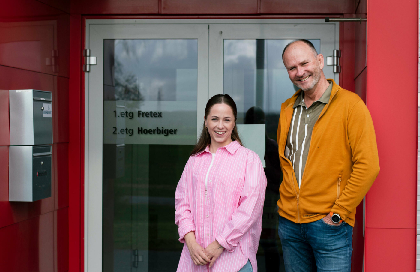 Foto: Kvinne og mann i 40-årene står utenfor en dør der det står "1 etg. Fretex". Hun har mørkt, langt hår og en rosa skjorte. Han er tynn i håret på toppen og har på seg en oransje jakke utenpå en beige og hvit skjorte.