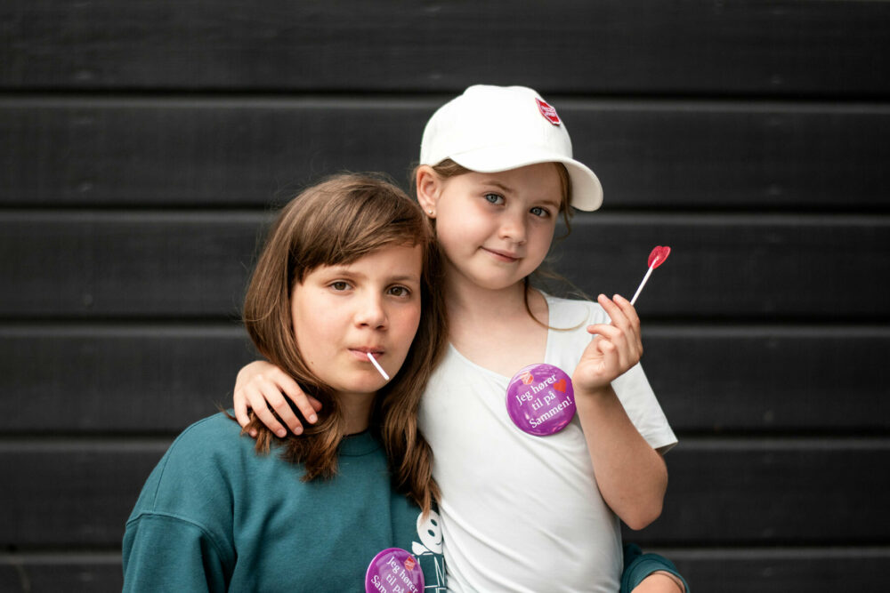 Foto: To jenter på barneskolen med hver sin kjærlighet på pinne. Begge har mørkt hår og en button der det står: "Jeg hører til på Sammen!"