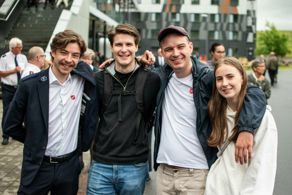 Foto: Fire ungdommer står på rekke og holder rundt hverandre. Tre er unge menn og den siste en tenåringsjente med mørkt, langt hår. En av gutta har på seg hvit skjorte og uniform fra Frelsesarmeen.