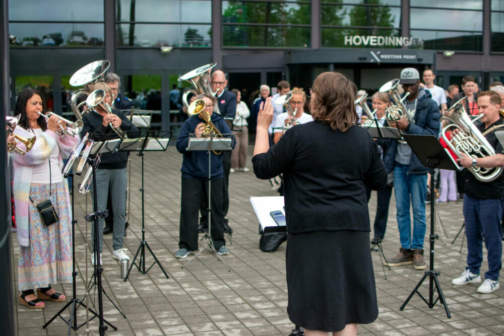 Foto: Hornorkester med en rekke brassinstrumneter står ute på en plass foran et bygg. Med ryggen til står en kvinnelig dirigent i mørkeblå klær.