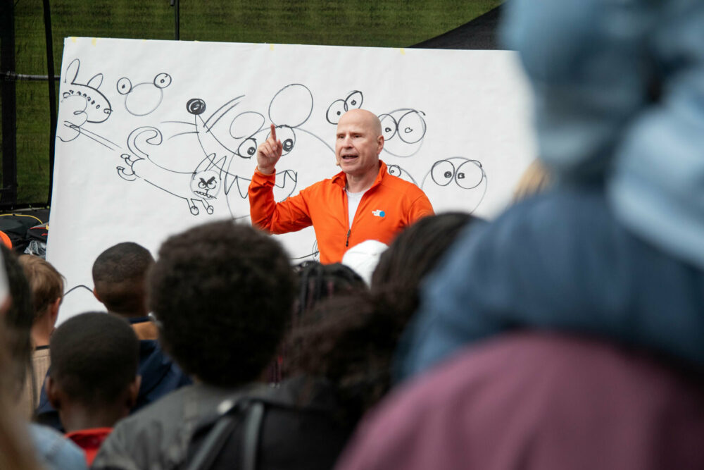 Foto: Mann uten hår i oransje fleecejakke står foran et stort, hvitt ark der han tegner figurer.