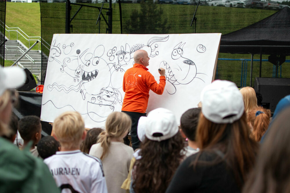 Foto: Mann uten hår i oransje fleecejakke står med ryggen til publikum og tegner på en hvit tavle.