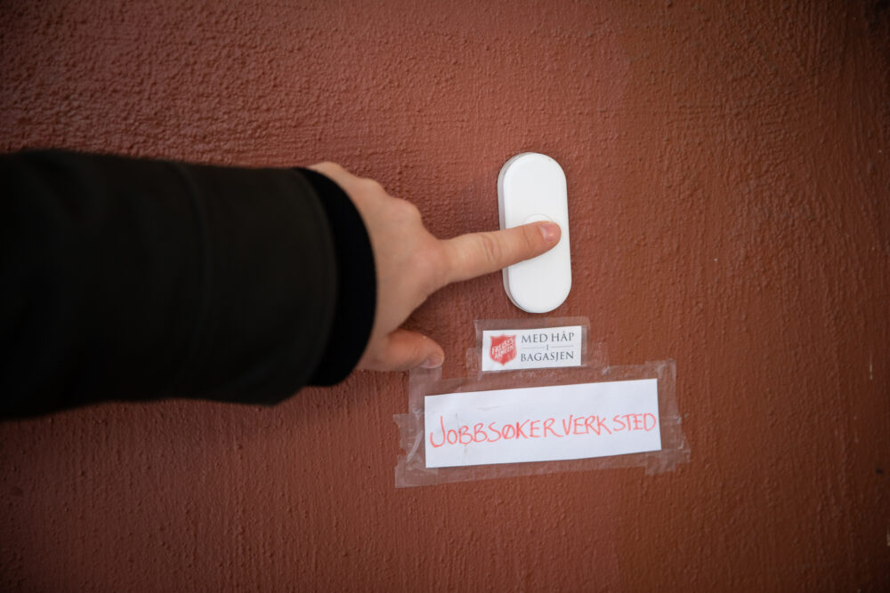 Foto: Hånd med pekefinger som trykker på en hvit ringeklokke. Det henger to lapper under, der det står "Med håp i bagasjen" og "Jobbsøkerverksted".