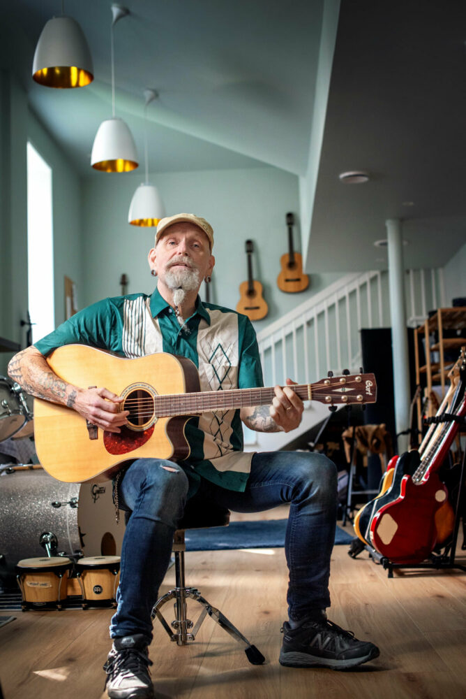 Foto: Mannen med skjedd og caps spiller gitar. Han sitter i et rom fylt med instrumenter.