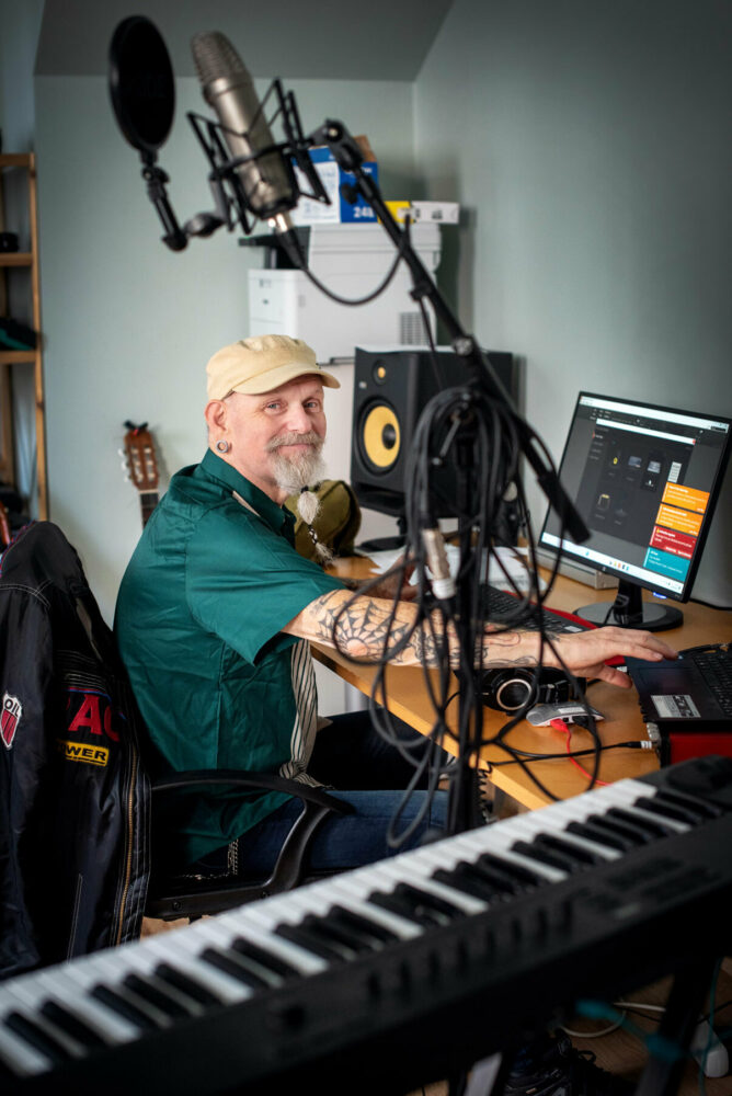 Foto: Mann med beige caps og grønn skjorte sitter ved en datamaskin i et musikkstudio. Han har langt skjegg og tatoveringer på begge armene.