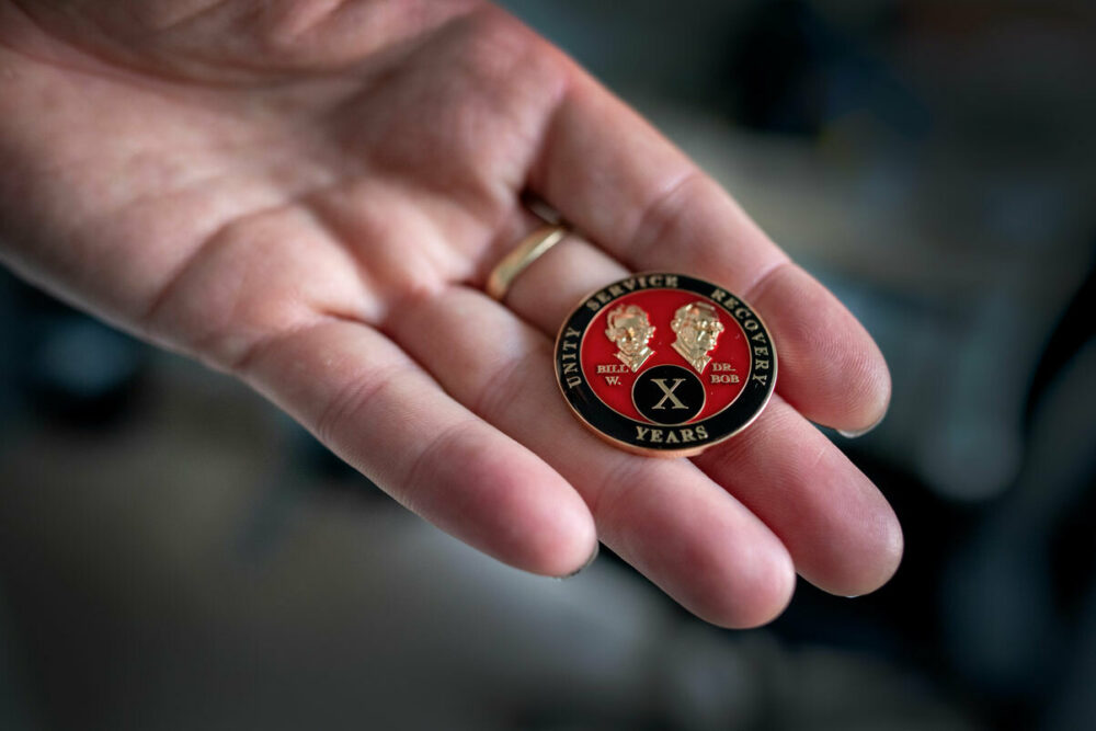 Foto: Mannshånd holder fram en pin i svart, rødt og gull med en X foran.