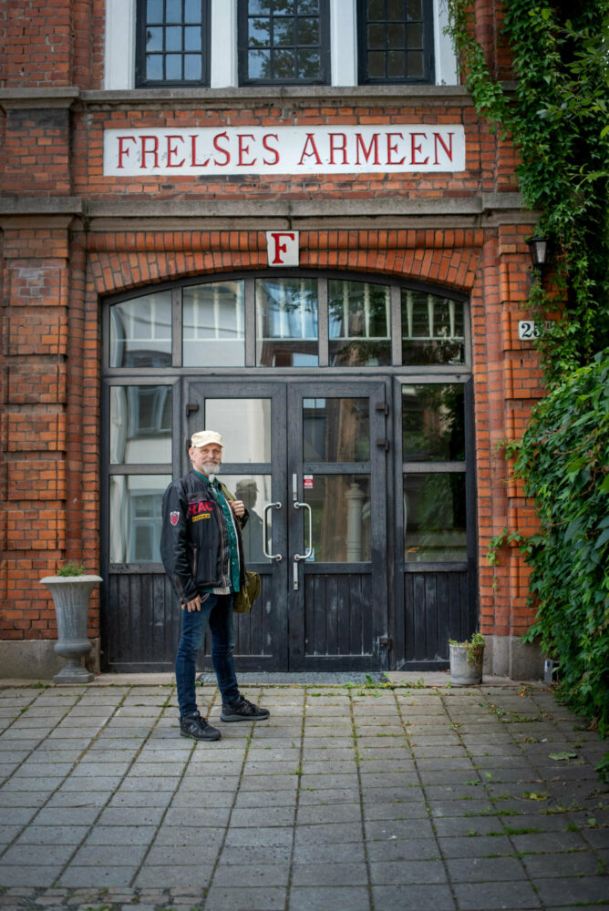 Foto: Mann med skinnjakke, grått skjegg og caps står utenfor en dobbel inngangsdør. Over døra til den røde mursteinsbygningen står det Frelsesarmeen murt inn med rødt på hvitt.