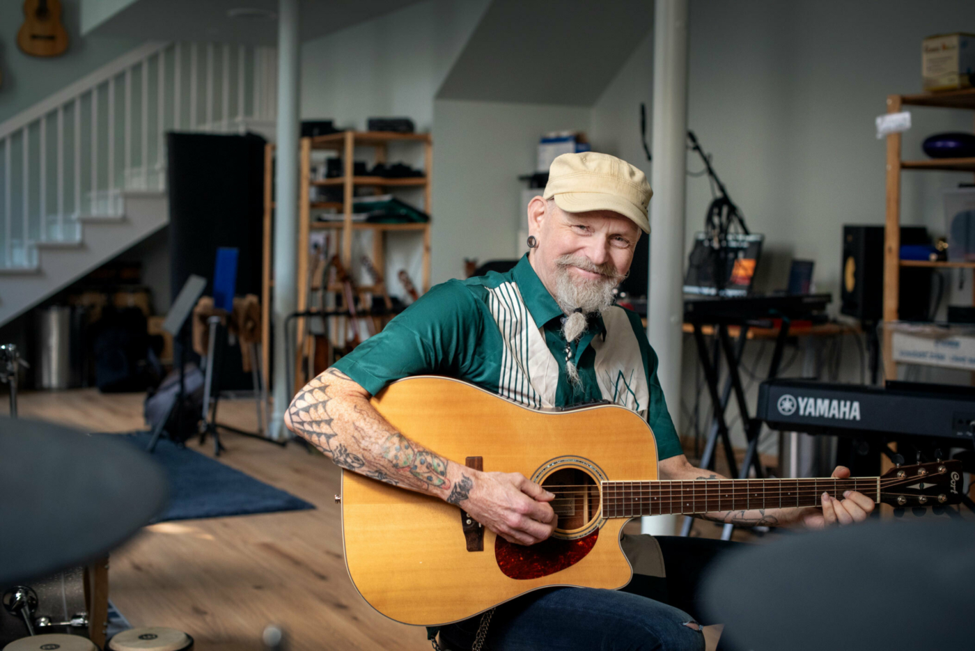 Foto: Mann i slutten av 50-årene med tatoveringer og skjegg sitter og spiller på gitar i et stort rom med mange instrumenter.