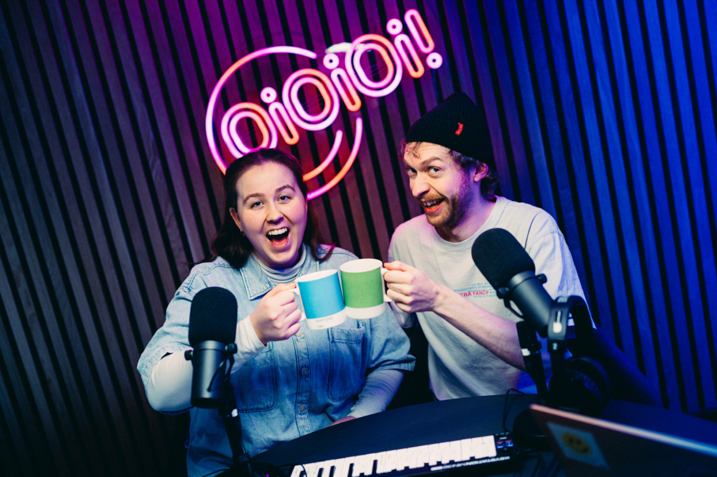 Foto: Kvinne og mann tidlig i 30-årene skåler med hver sin kopp i blått og grønt. De sitter i et podkast-studio og bak på veggen er det et rosa neonskilt der det står navnet: oioioi!