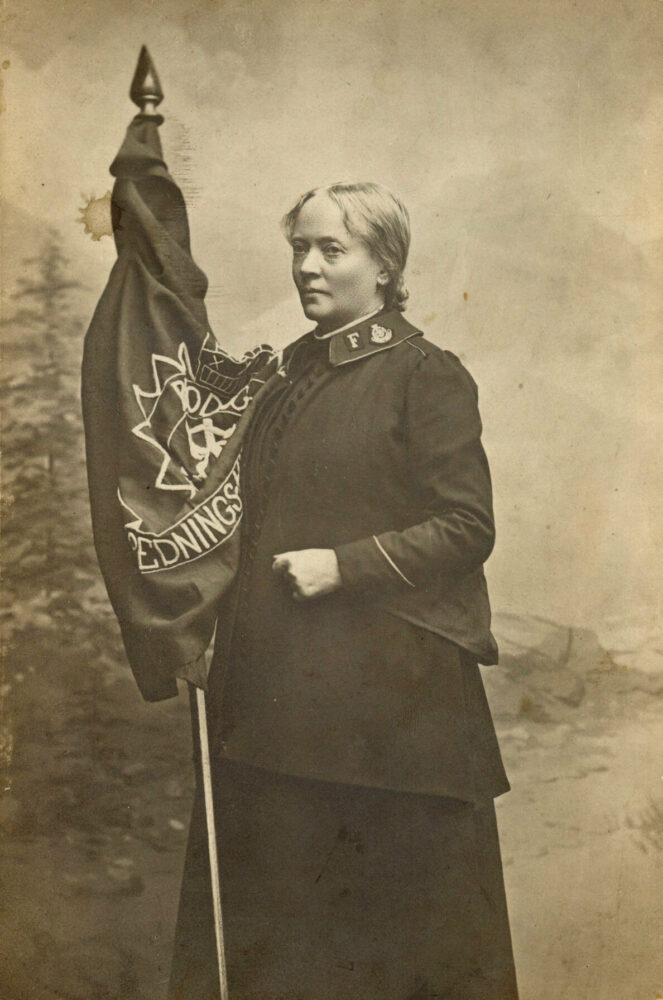 Foto: Gammelt i brungule toner viser en kvinne i uniform fra Frelsesarmeen som står med er flagg fra Frelsesarmeen.