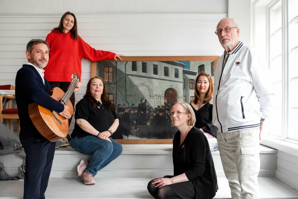 Foto: Fire kvinner og to menn, den ene med en gitar, står rundt fotografiet av en kvinne på en tønne utendørs i en bakgård på slutten av 1800-tallet.