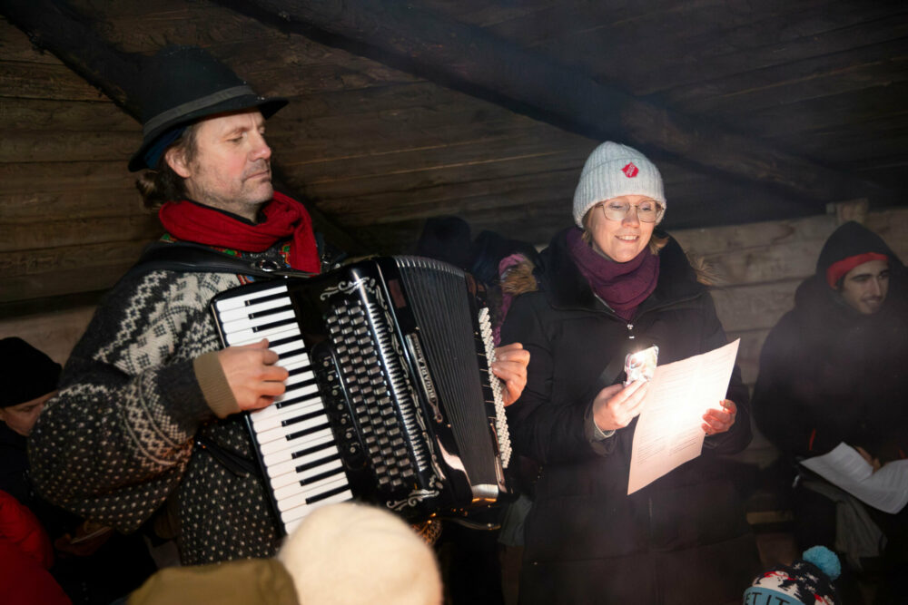 Foto: Mann i strikkengenser, rødt skjerf og hatt spiller trekkspill. Ved siden av ham sitter en dame med mobil og lyser opp et ark med sangtekster med mobilen sin. Det sitter flere folk rundt i en lavvo i tre.