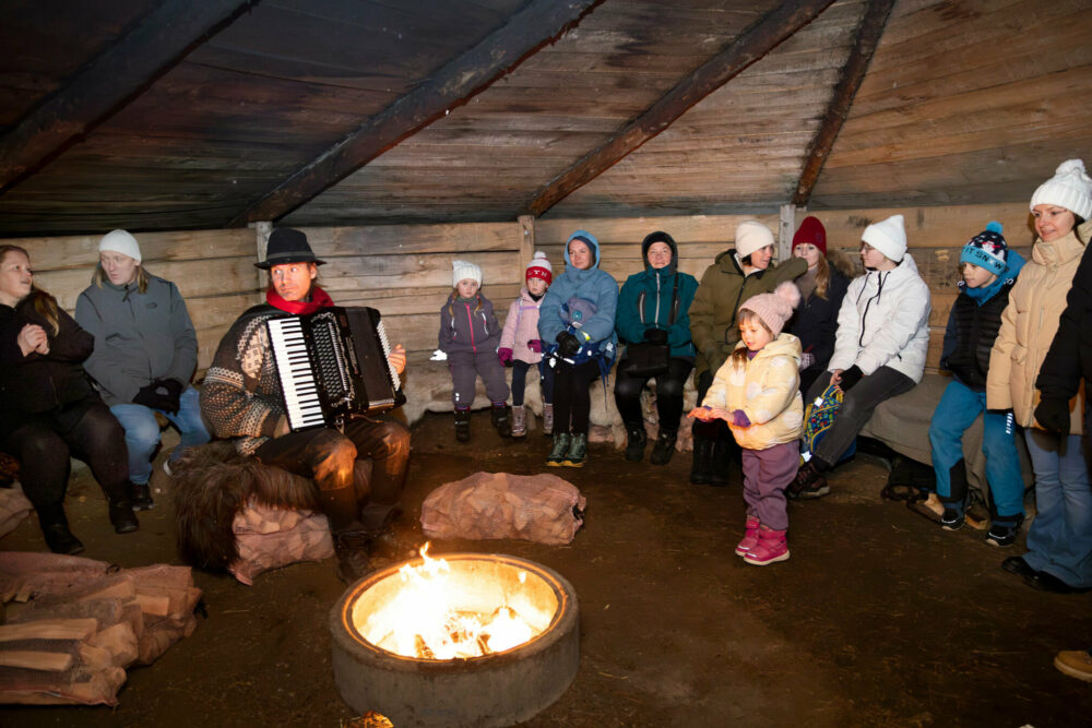 Foto: Rundt 15 små og store mennesker inni en lavvo av tre. I midten er det en bålpanne, og en mann spiller trekkspill.