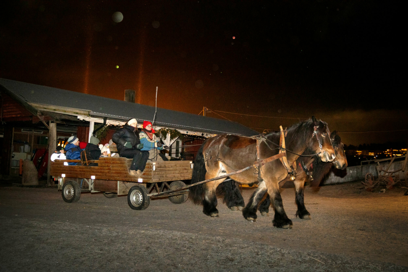 Foto: To brune hester drar en kjerre full av barn og voksne gjennom mørket. Det er litt snø på bakken og en gårdsbygning i bakgrunnen.