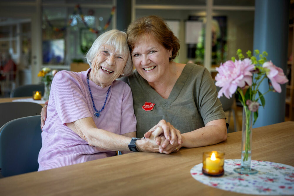 Foto: Kvinne i 50-årene med mørkt hår sitter ved et kafebord sammen med en gråhåret eldre kvinne. De sitter tett, smiler veldig og holder hender.