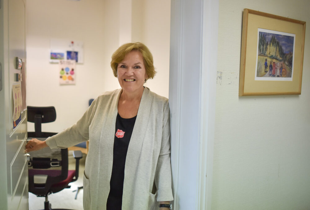 Foto: Kvinne med mørkt hår i 50-årene står i en døråpning inn til et kontor. Hun har på seg svart T-skjorte med logo fra Frelsesarmeen og en beige strikkejakke.