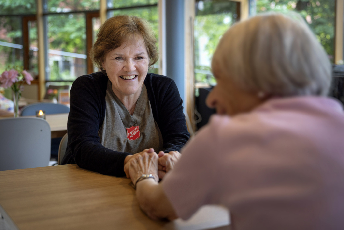 Foto: Smilende kvinne i 50-årene med mørkt hår holder hendene til en gammel dame med grått hår. De sitter på hver sin side av et bord og strekker hendene over det. Den eldre damen ar ryggen til.