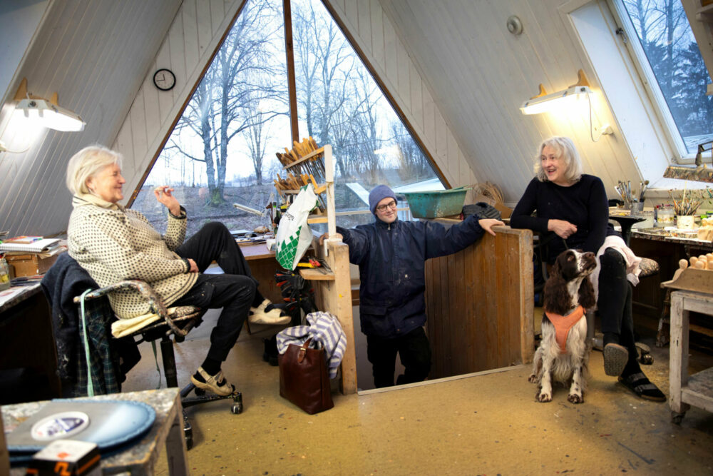 Foto: To eldre kvinner sitter på hver sin side på et loft med spist tak og av et loft med trekantet vindu i enden. Mellom dem kommer det en trapp opp, og der står en yngre mann med lue og briller.