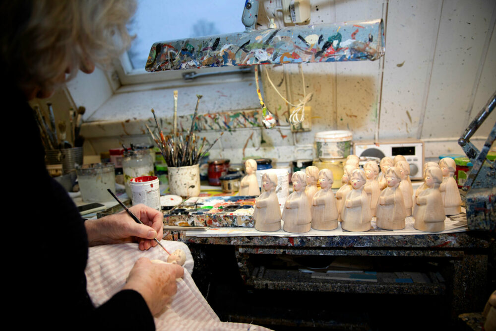 Foto: Kvinne i svart genser med grått hår maler på et helt brett med engler. På bordet står en rekke pensler.