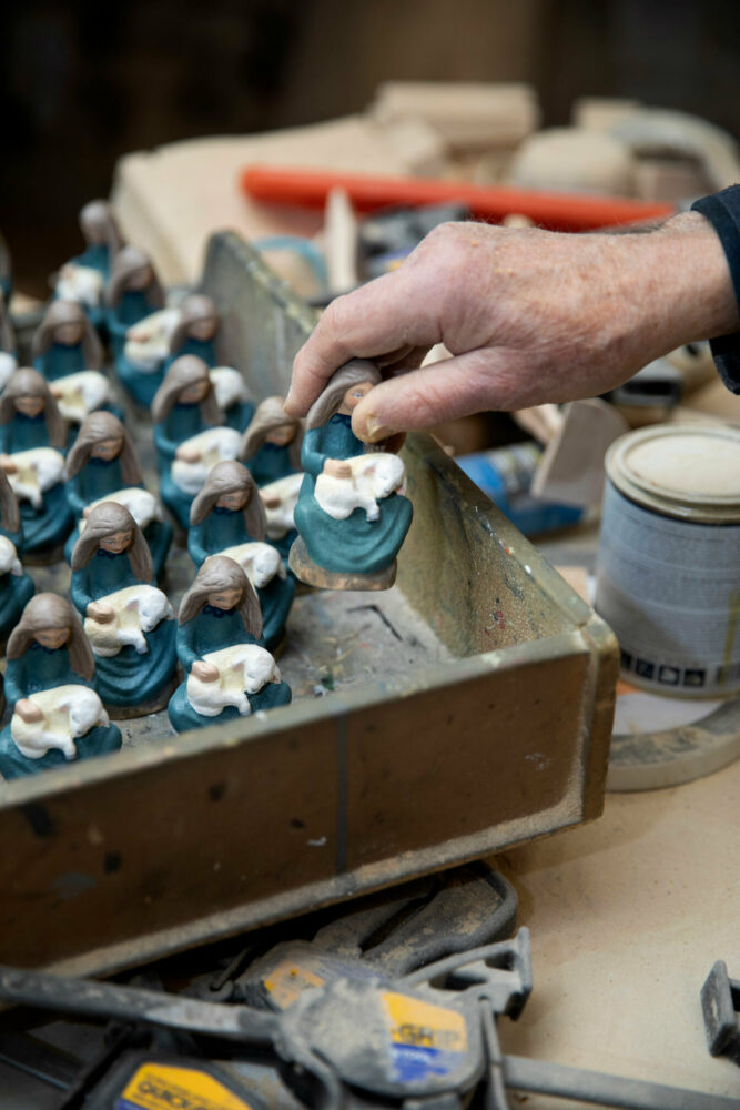 Foto: En rekke like figurer står til tørk i en boks. De viser en kvinne med langt, blondt hår som sitter og holder et hvitt lam i armene sine.