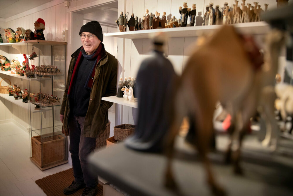 Foto: Mann i 70-årene med briller står i yttertøyet i en hvitmalt butikk. Der er det hylle på hylle med figurer i tre, alt fra bibelsk figurer til troll.