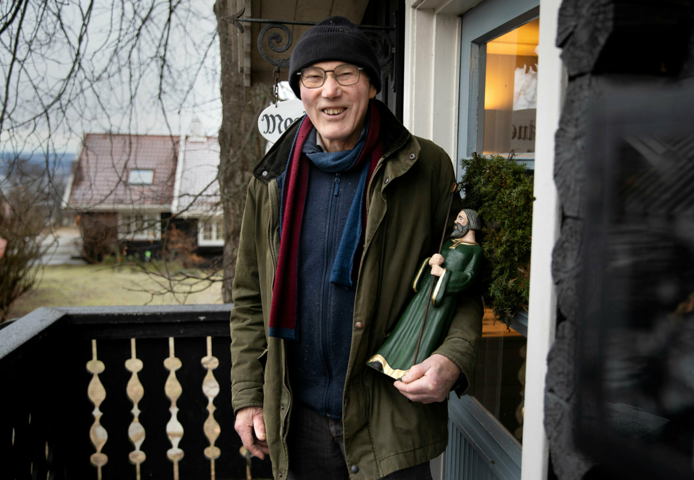 Foto: Eldre mann i mosegrønn jakke står ved inngangsdøra til et verksted. Han har briller og lue på. I venstre armkrok står en statue av en mann i grønn kappe med en stav.