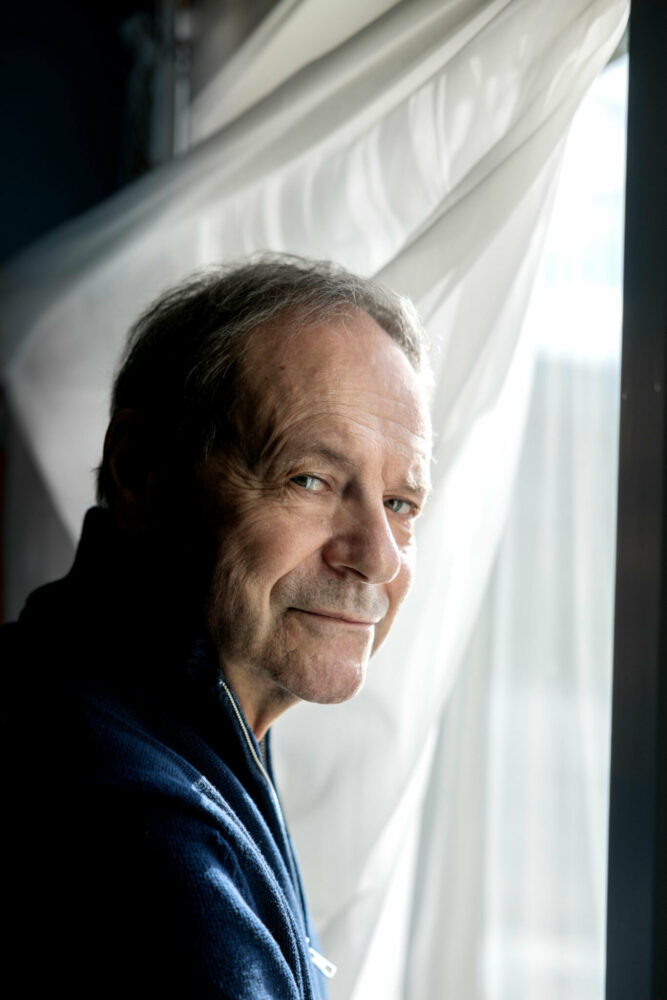 Foto: Mannen i 60-årene med mørkeblå genser ser i kamera. Han står foran en hvit gardin i et vindu.