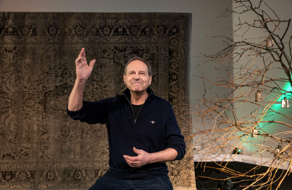 Foto: Mann sitter på en barkrakk på en scene og holder høyre arm opp i luften og venstre på magen. I bakgrunnen henger et stort persisklignende teppe i brune og beige toner. På høyre siden av ham stikker det inn kvister hvor det henger en rekke bjeller.