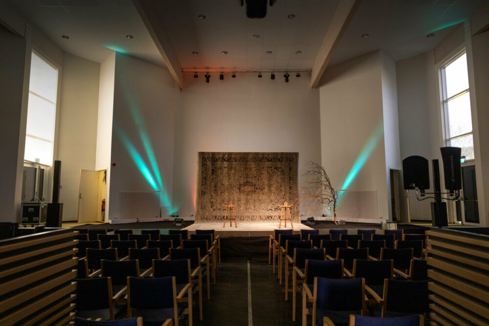 Foto: Av en stor sal med mange stoler og en scene i enden. Der henger det et teppe i enden. Her er det grønne lyskastere oppover veggen.