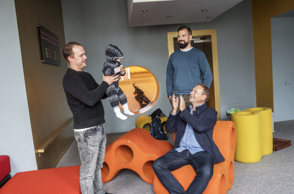 Foto: Far i 60-årene med to sønner i 30-årene og et barnebarn som er baby. De er i et fargerikt rom med mange puffer og andre former.