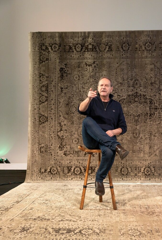 Foto: Mann i 60-årene med mørkeblå genser sitter på en krakk på scenen. Han peker rett fram mot salen med sin høyre hånd, armen er utstrakt.