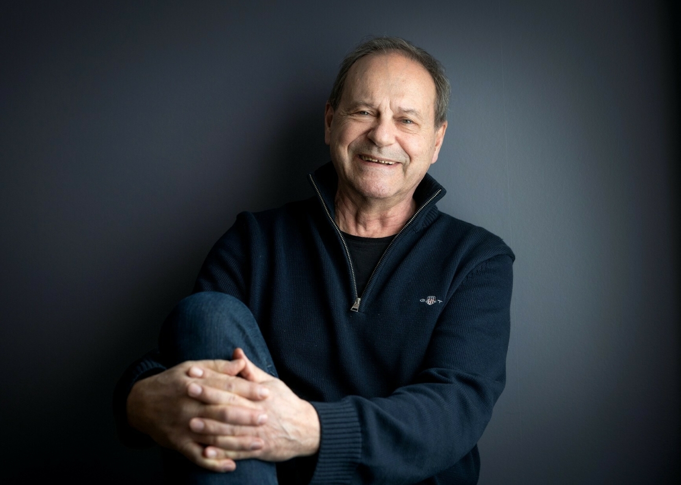 Foto: Mann i mørkeblå genser sitter avslappet inn mot en mørkeblå vegg. Han holder hendene foldet foran det ene kneet sitt og smiler skjevt til kamera.