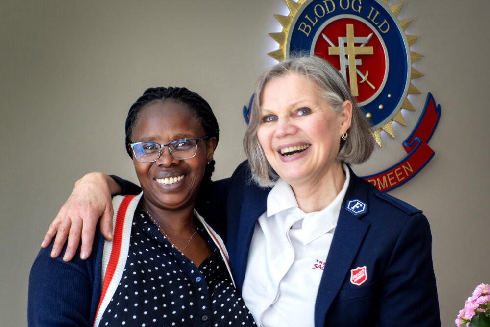 Foto: Hvit kvinne i 60-årene står med armen rundt en svart kvinne med briller. De står under et skilt fra Frelsesarmeen der det står blod og ild.