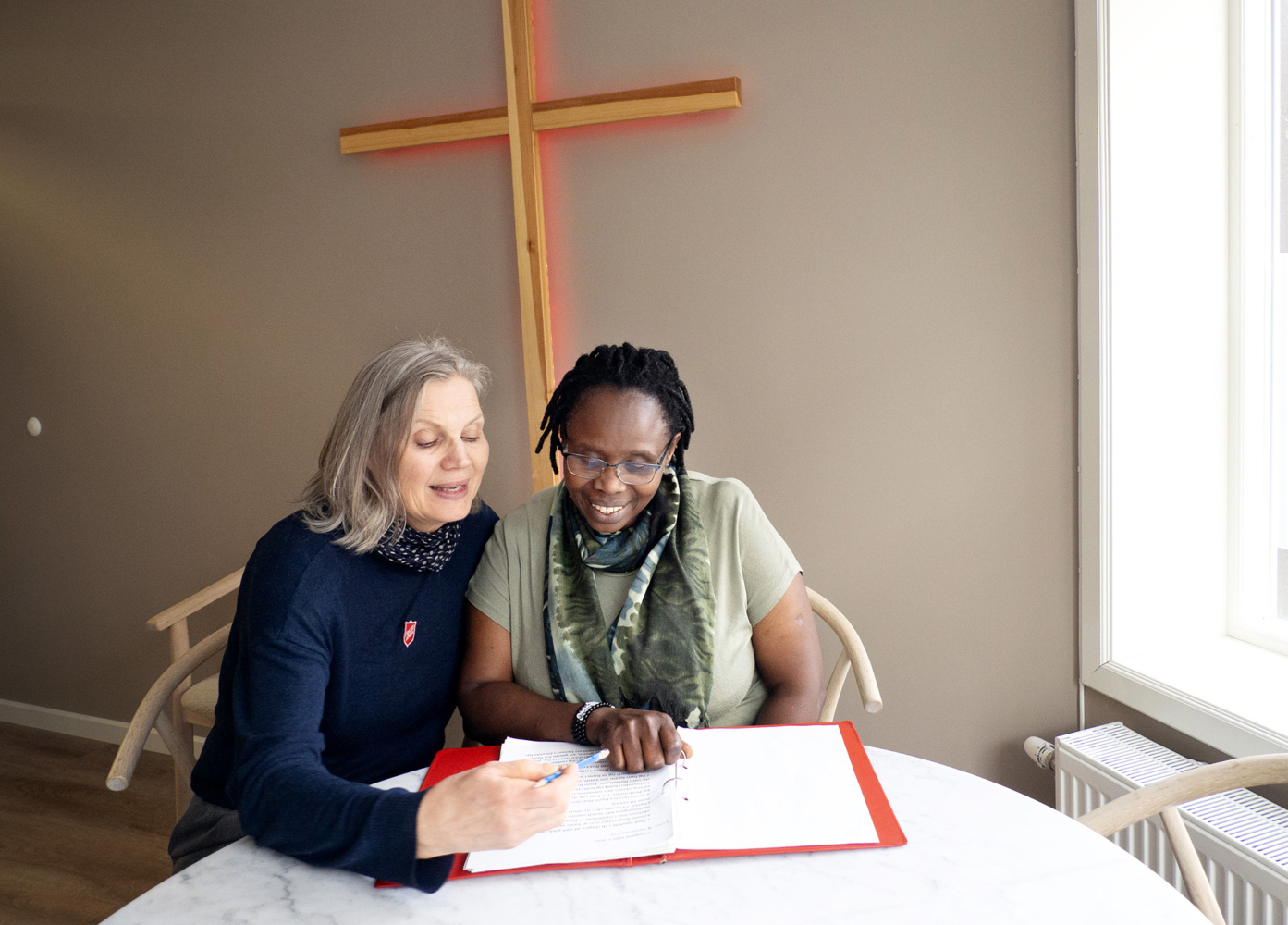 Foto: To kvinner sitter ved et bord og ser ned i en rød perm med hvite ark og svart tekst. Den ene er hvit og den andre svart. På veggen bak henger et kors med lys bak.