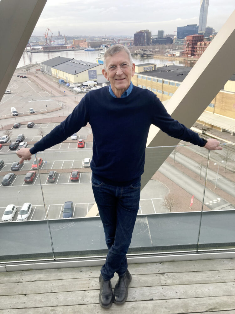 Tomas Sjödin står med ansiktet mot kamera. Bak ham er det et heldekkende vindu som viser utsikten fra toppetasjen i Frihamnskyrkan i Gøteborg.