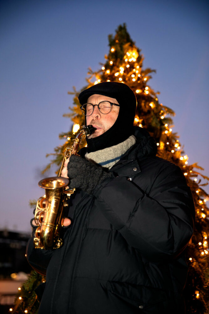 Foto: Mann i vinterklær står foran en tent julegran og spiller på en gyllen saksofon.