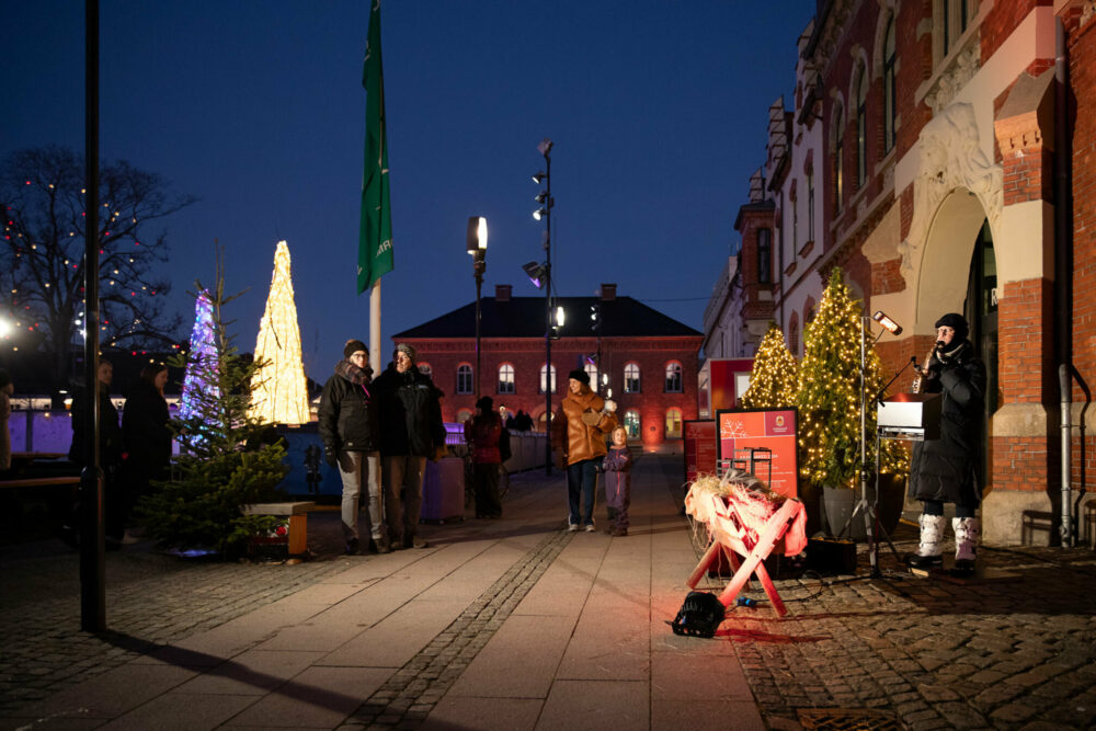 Foto: Mann med saksofon står foran et gammelt rødt bygg på et torg. Folk står rundt ham. Det står også flere juletrær med lys rund på området også.
