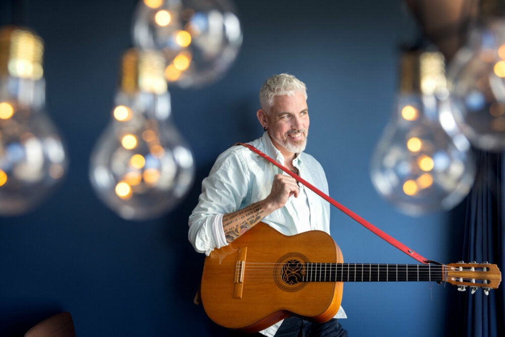 Foto: Mann med grått hår og gitar i tre med rødt bånd. Rundt ham er det en rekke lyspærer.