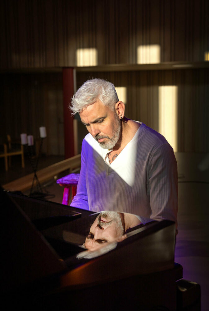Foto: Mann med grått hår og skjegg sitter ved pianoet. Ansiktet hans reflekteres i den svarte, blanke overflaten.