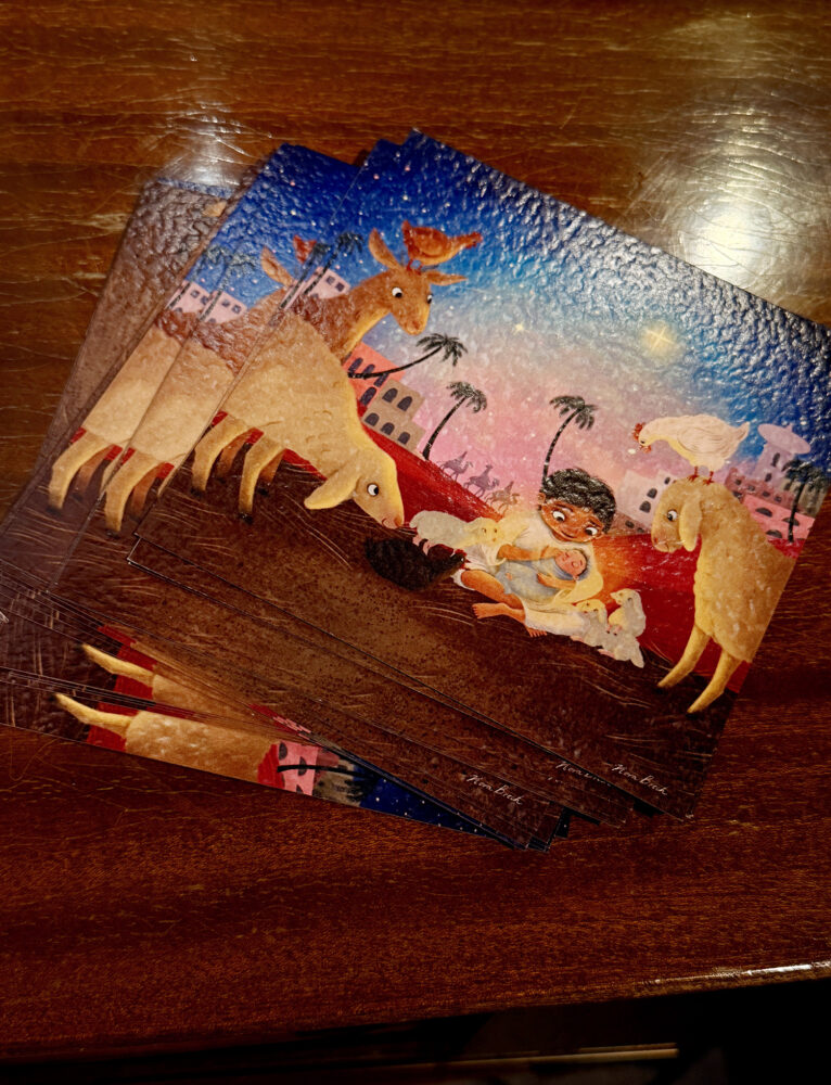 Foto: En liten bunke med julekort ligger på et bord. Det er en fargerik tegning av en gutt som holder en liten baby.