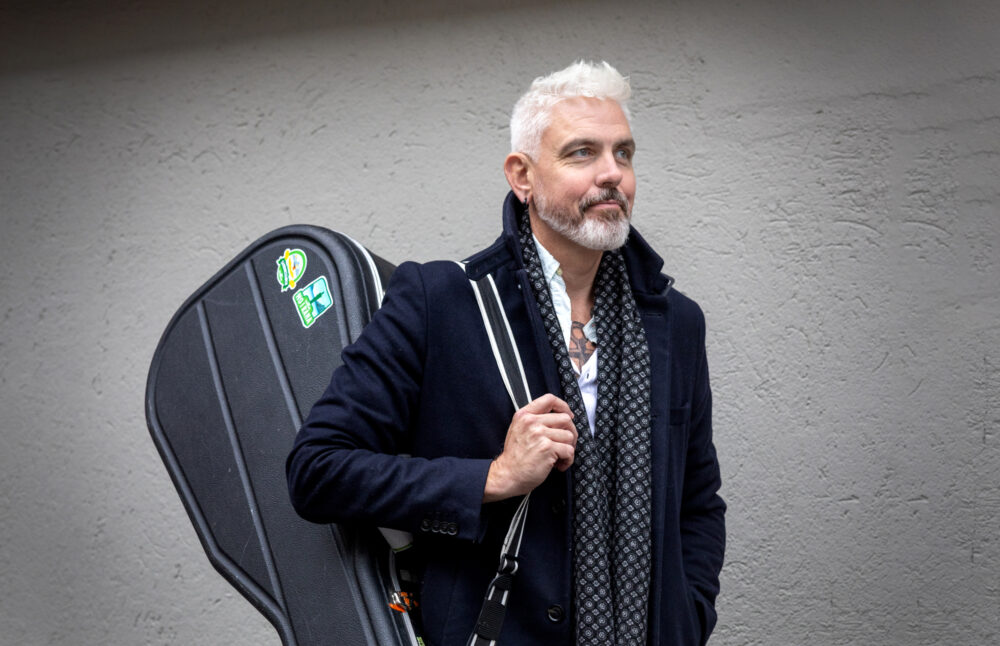 Foto: Mann med grått hår i svart frakk. Han har en svart gitarkasse hengende på høyre skulder.