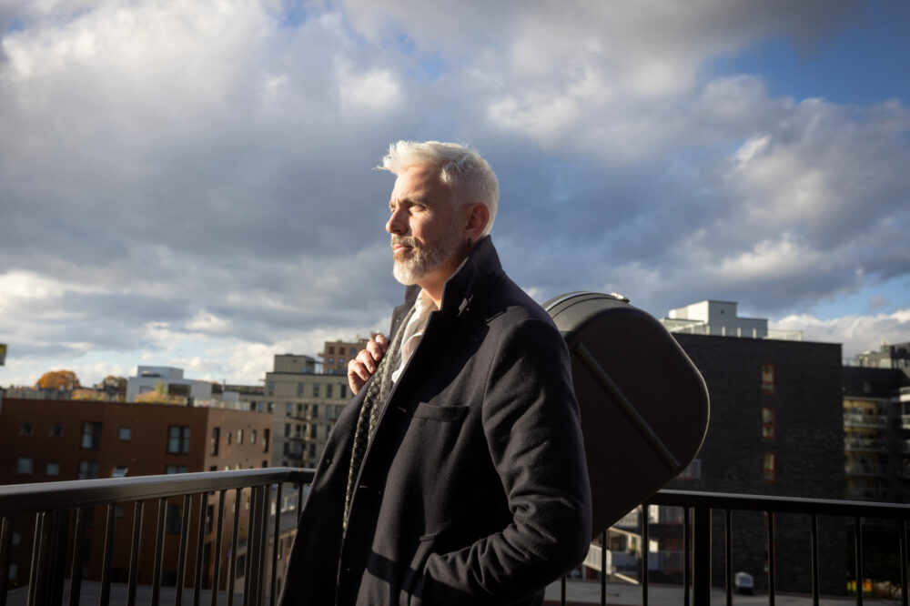 Foto: Mann med grått hår og skjegg har på seg frakk og gitaren over skulderen. Han står på en terrasse og det er skyer på himmelen, men noen solstråler kommer igjennom og det er en flik av blå himmel her og der.