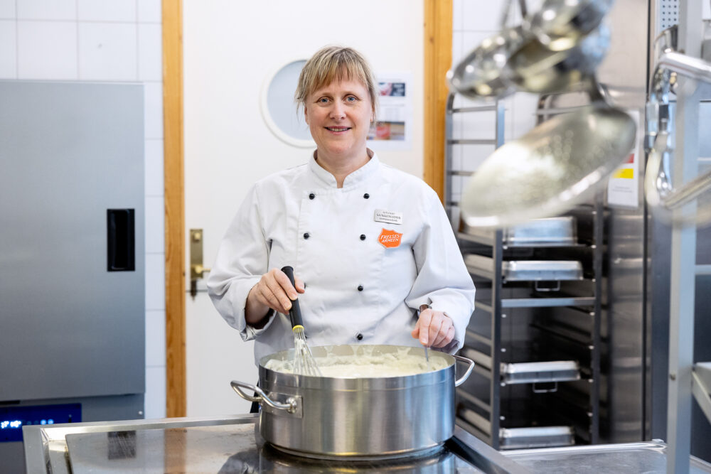 Kvinne i hvit kokkeuniform rører i en diger gryte.