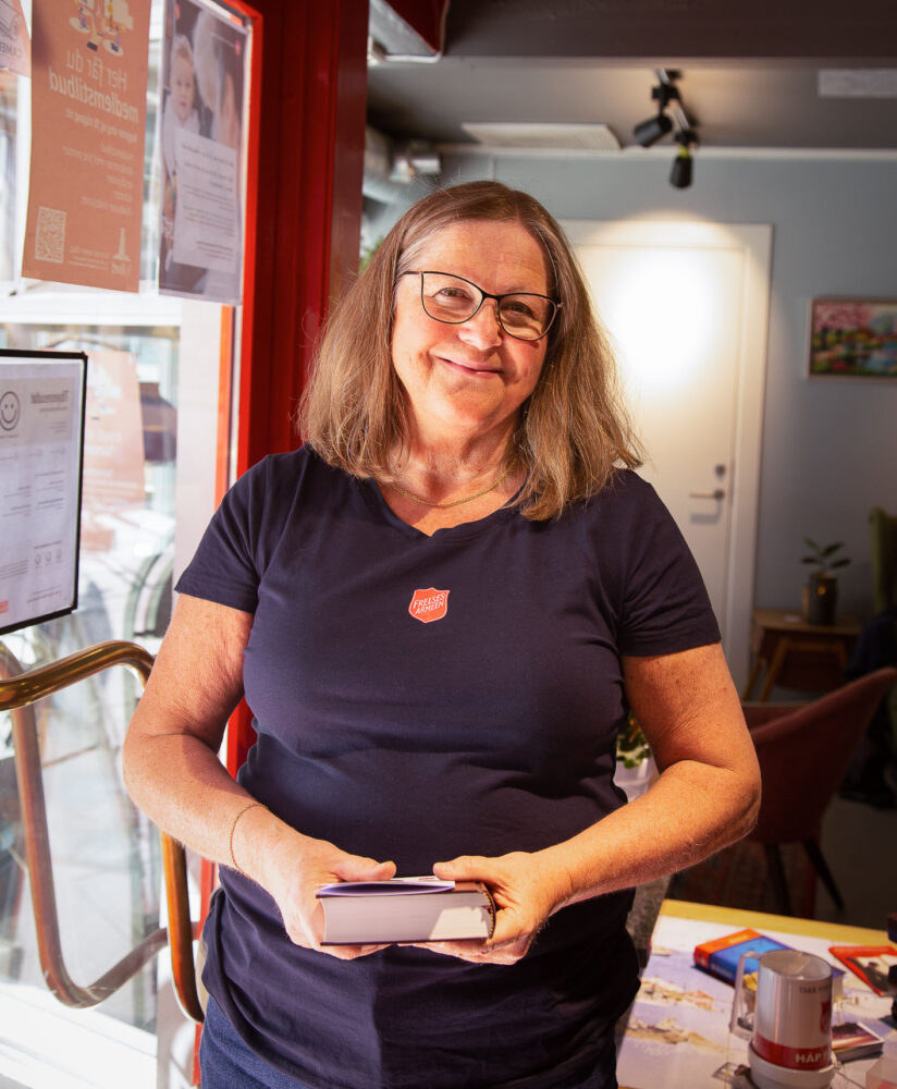 Dame med blå Frelsesarmee-T-skjorte står og smiler med en bok i hendene.