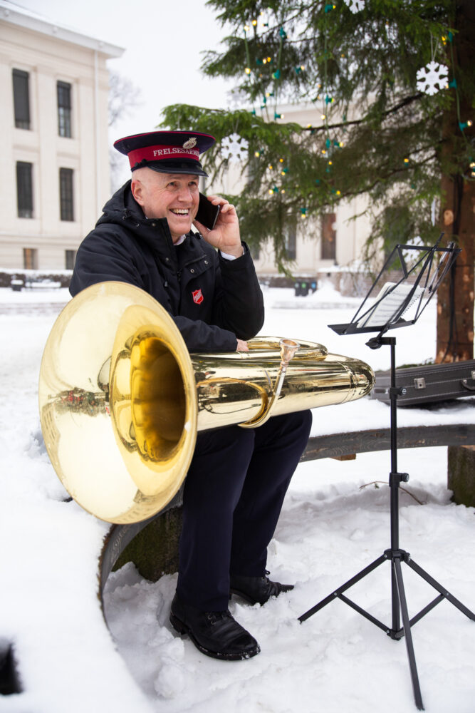 Mann mørk uniform og hatt med stor tuba på fanget. Snakker i mobilen.