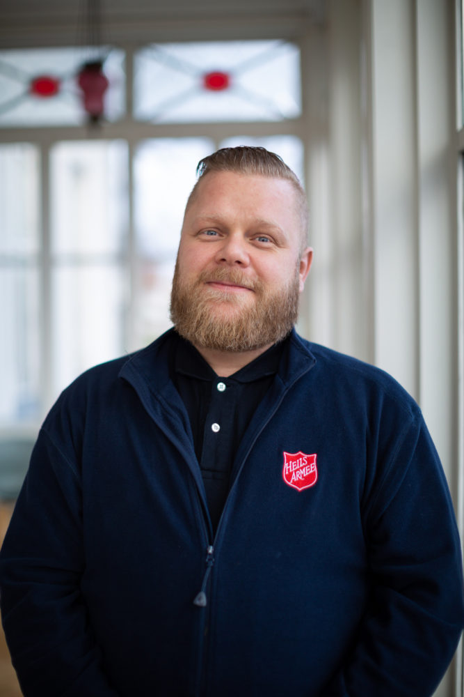 Kadett Sigurður Ívar Sölvason står på glassverandaen på Frelsesarmeens offiserskole på Jeløy. Han har på seg en en mørkeblå genser med Frelsesarmee skjldet på brystet.