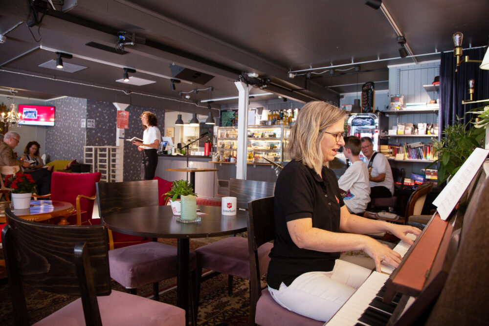 Kvinne i uniform sitter vet pianoet. I bakgrunnen ser man gjester i kafeen.